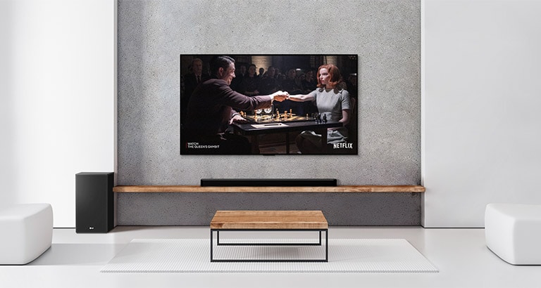 Um conjunto de 2 alto-falantes traseiros, um subwoofer e uma barra de som, bem como a TV, estão localizados em uma sala de estar branca. Uma mulher e um homem jogam xadrez na tela da televisão.