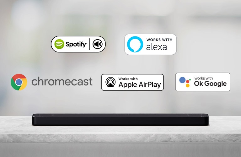 Há uma barra de som localizada em uma prateleira cinza e logotipos de plataformas de IA, na ordem de Spotify, Alexa, Chromecast, Apple Airplay e OK Google da esquerda para a direita.