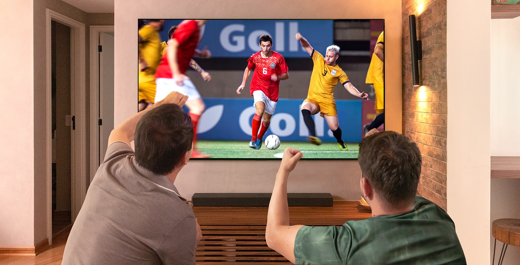 Dois amigos torcendo com os braços levantados, punhos cerrados para a partida de futebol jogada na TV LG OLED na sala de estar.