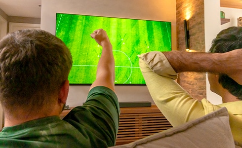 Dois amigos torcendo, um com o punho alto e o outro segurando a cabeça. Eles estão olhando para a tela do campo de futebol na TV LG OLED.