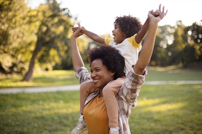 Mãe e filha afro-americanas em pé num parque. A menina está sentada nos ombros da mãe