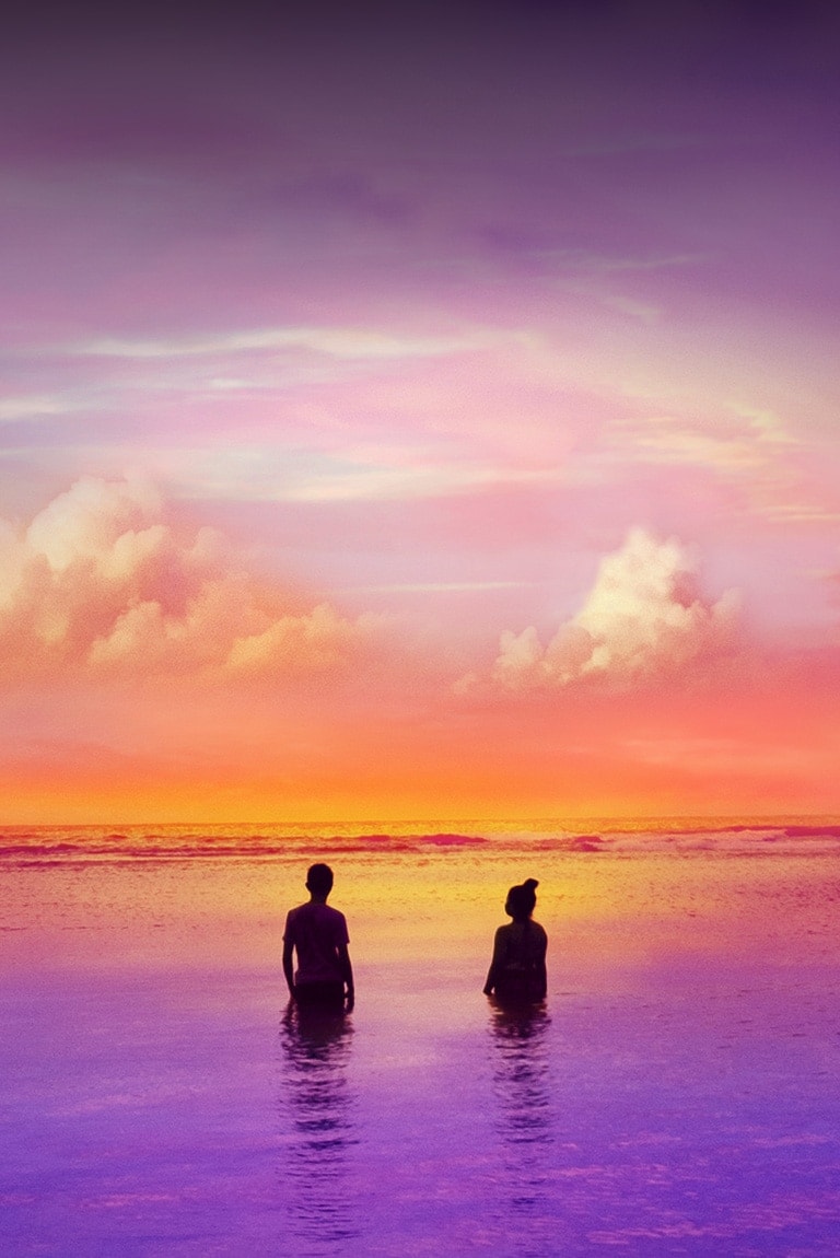 Duas pessoas estão de pé dentro do mar, que tem coloração ligeiramente púrpura. É um fim de tarde.