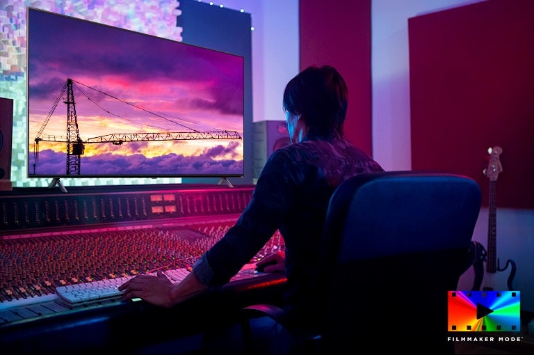 Um diretor de cinema está editando algo em um grande monitor de TV. A tela da TV mostra um guindaste de torre num céu arroxeado. O logotipo FILMMAKER Mode está colocado no canto inferior direito