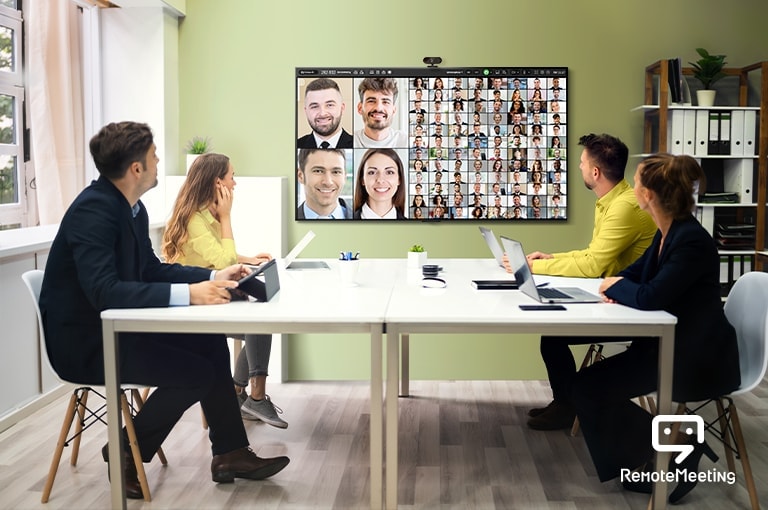 Quatro pessoas estão sentadas em uma sala, fazendo uma teleconferência pela TV. A tela da TV mostra os rostos das pessoas que participam da reunião