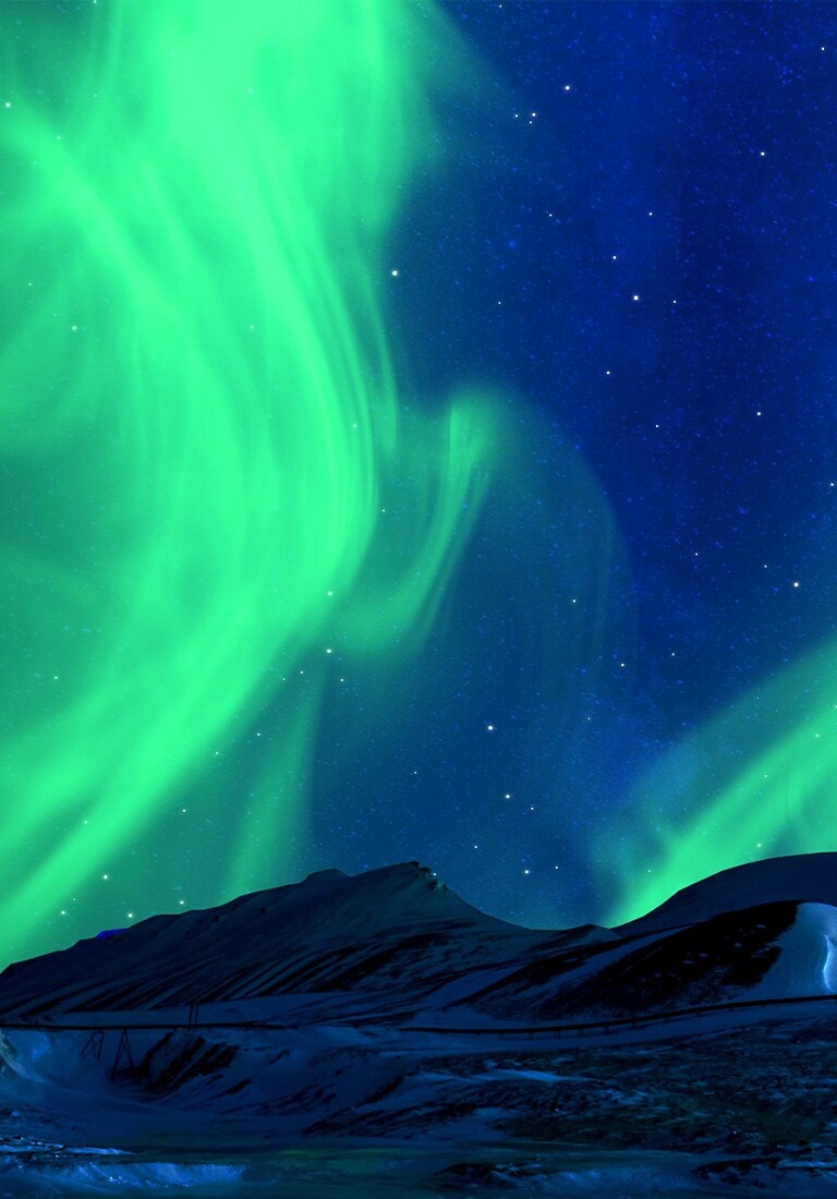 Paisagem de inverno composta de montanhas nevadas e céu noturno estrelado com aurora boreal esverdeada ao fundo.
