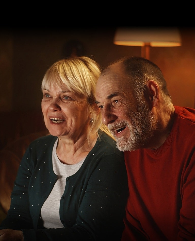 Um casal de idosos está sentado em um sofá, sorrindo e olhando para a tela.