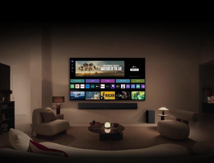 A close-up of an LG TV screen showing the buttons Home Office, Game, and Music over a banner for Masters of the Air zooms out to show the TV mounted on a wall in a living room. The following logos are displayed on the TV screen in the image: LG Channels, Netflix, Prime Video, Disney TV, Apple TV, YouTube, Spotify, Twitch, GeForce Now and Udemy.