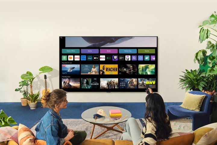 A whale floating above the ocean with a woman in the ocean are shown. A home screen appears from below. As the scene transitions, it shows the image in a large LG TV on the wall. Two women sit in a cozy and neutral living room filled with plants, and a guitar. One woman points the remote at the TV, which shows a range of apps and recommended TV shows on the home screen. 