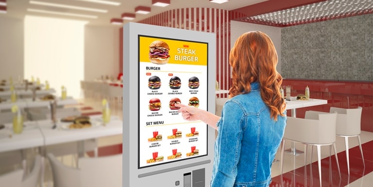 A woman is touching a kiosk to select a hamburger from the menu.