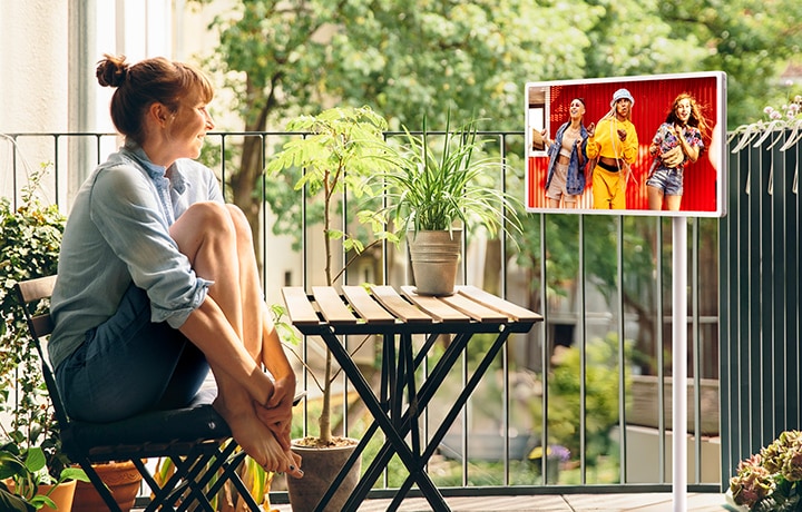 A woman sitting in her outside balcony, watching content on an LG StanbyME 2 on the rollable, adjustable stand.