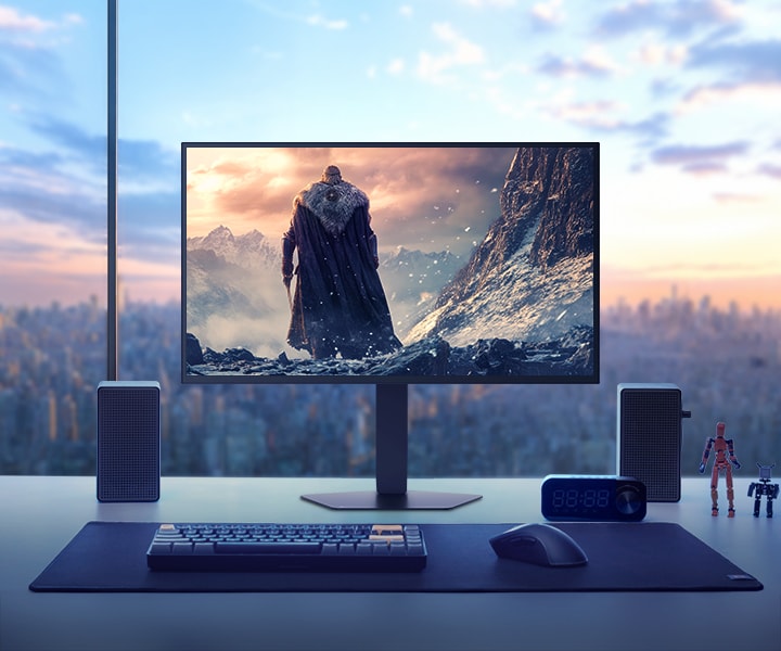 LG UltraGear 27GX790B monitor placed on a desk overlooking a cityscape at sunset, displaying a fantasy game scene with a cloaked character standing in a snowy mountain environment. Surrounding the monitor are speakers, a keyboard, a clock, small figurines, and a mug