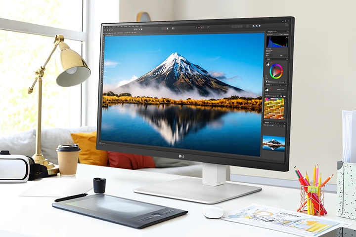 En el escritorio, hay un monitor ultrafino que muestra trabajos de Photoshop. Junto al monitor, hay una lámpara de escritorio, un mouse, papel y lápices de colores.