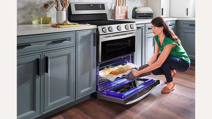 Woman in green top placing a baking tray with a roast into an open oven in a modern kitchen with grey cabinets.