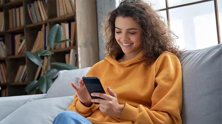 The woman is sitting on the sofa and using her cell phone