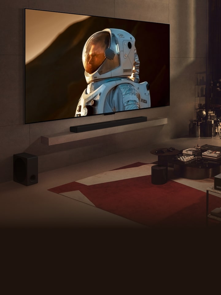 Un LG TV de très grande taille fixé au mur d’un salon confortable et affichant un gros plan d’astronaute occupe la majeure partie du mur. Un homme est assis sur un canapé et regarde la télévision en compagnie d’un chien. Un paysage urbain nocturne se dessine derrière la fenêtre.