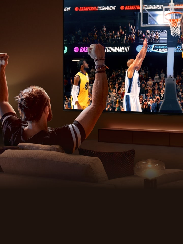 Un homme est assis sur un canapé devant un LG TV très grand qui affiche un match de basket, les bras en l’air comme pour applaudir.