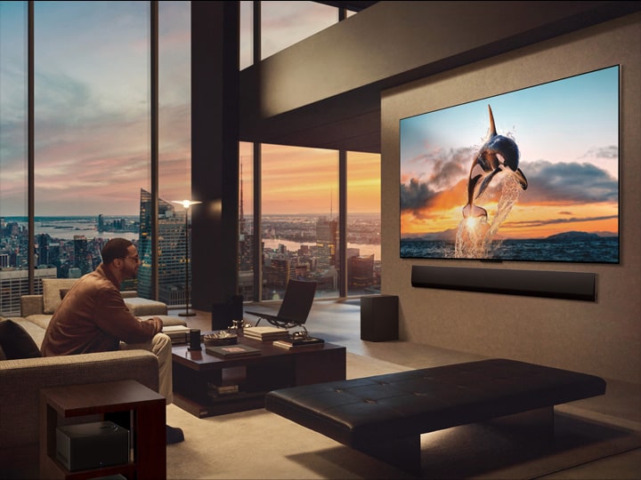 Geräumiges Wohnzimmer mit Blick auf eine Stadtlandschaft. Der LG OLED TV ist mit einer LG Soundbar an der Wand befestigt. Das flächenbündige Design des Fernsehers ermöglicht eine bündige Montage an der Wand und fügt sich gut in den Raum ein. Die Zero Connect Box ist zu sehen, aber unauffällig aufgestellt.