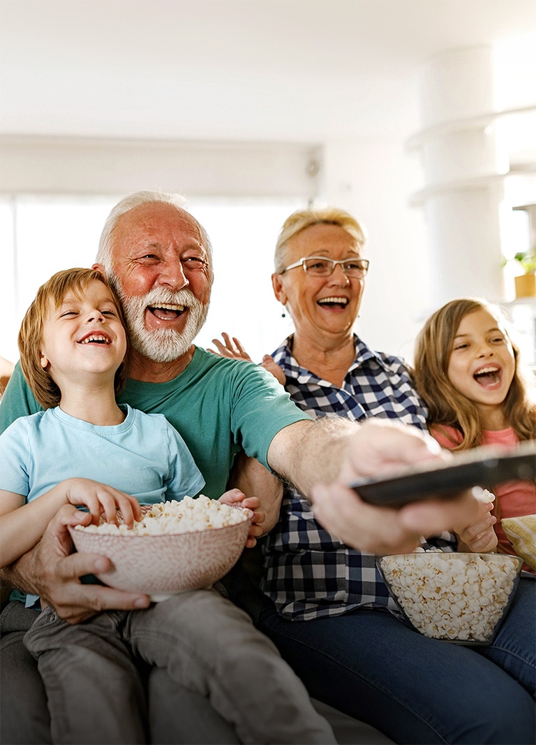 Eine Familie sitzt lachend zusammen, während der Großvater die Fernbedienung nach außen richtet.