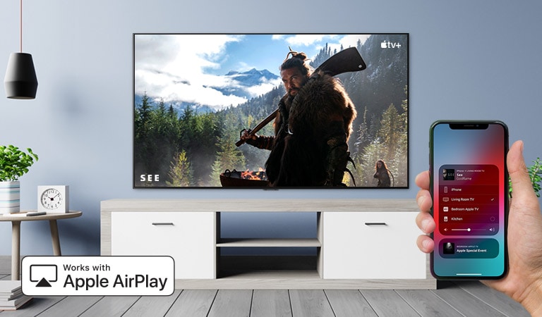 Nahaufnahme einer Hand, die den TV über ein iPhone steuert. Auf dem Fernseher werden Inhalte von Apple TV+ angezeigt.