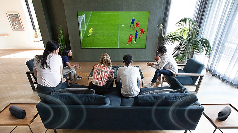 5 Personen, die vor einem wandmontierten Flachbildfernseher sitzen und sich ein Fussballspiel ansehen.