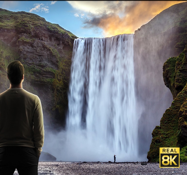 Ein Mann steht vor einem imposanten Wasserfall, der die Klippen hinunterstürzt. Aus der Szene wird herausgezoomt, um den Wasserfall als Bild auf einem wandmontierten Fernseher zu zeigen.
