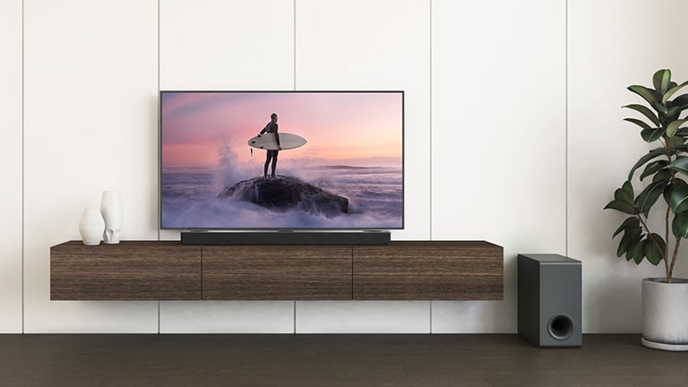 Un téléviseur LG et une barre de son LG sont placés sur une étagère marron et le caisson de basses est installé au sol. L’écran du téléviseur montre un surfeur debout sur le rocher.