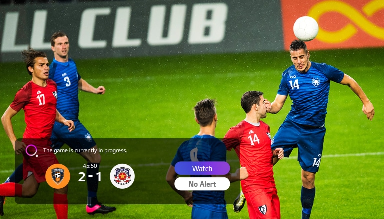 Un couple assis sur un canapé regarde un match de football sur un téléviseur qui affiche une notification d’un autre match en bas à gauche.