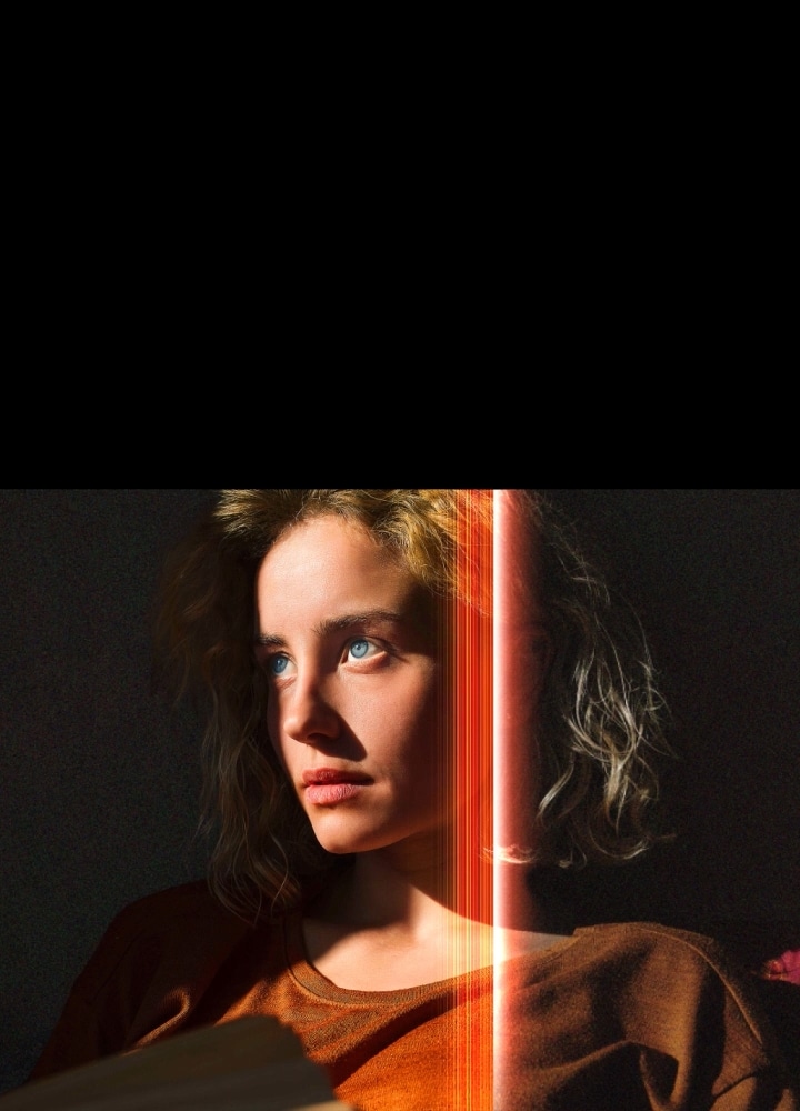 Una mujer con penetrantes ojos azules y una blusa naranja quemada en un espacio oscuro. Líneas rojas que representan los refinamientos de IA cubren parte de su rostro, que se ve vivo y detallado, mientras que el resto de la imagen se ve apagada. 