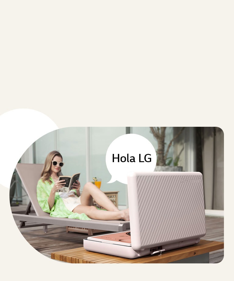 Vista trasera del LG StanbyME Go, y está colocado justo en frente de la mesa del patio. Una mujer se relaja en la silla de playa, controla la pantalla con su voz. Para ilustrar esto, se muestra un bocadillo con el texto Hola, LG en el lado derecho de ella.