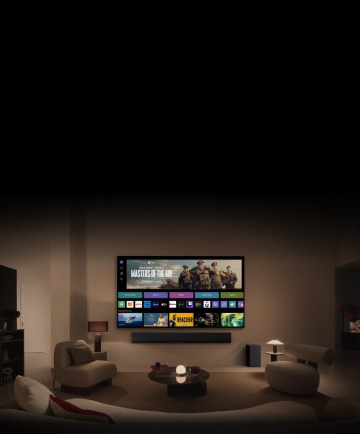 Un primer plano de la pantalla de un TV LG que muestra los botones Home Office, Game y Music sobre un banner de Masters of the Air se aleja para mostrar el TV montado en la pared de un salón. En la imagen aparecen los siguientes logotipos en la pantalla del TV: LG Channels, Netflix, Prime Video, Disney TV, Apple TV, YouTube, Spotify, Twitch, GeForce Now y Udemy.