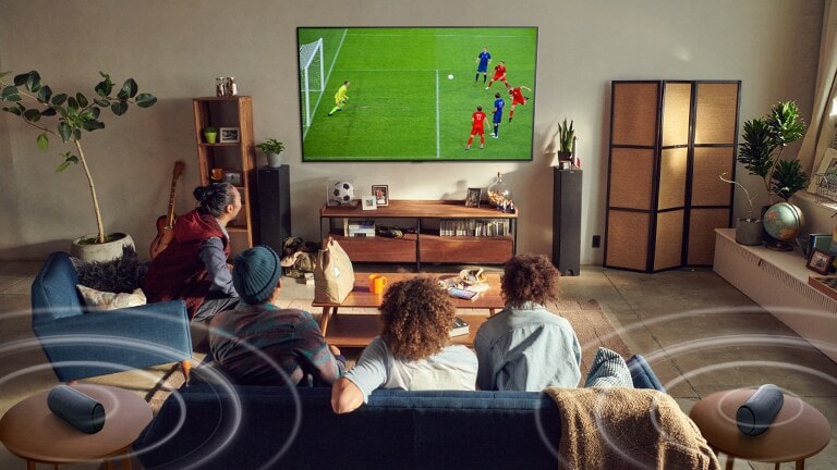 Cinco personas sentadas en el sofá miran un televisor de pantalla grande que muestra un juego de fútbol con altavoces Bluetooth que emiten un sonido envolvente.