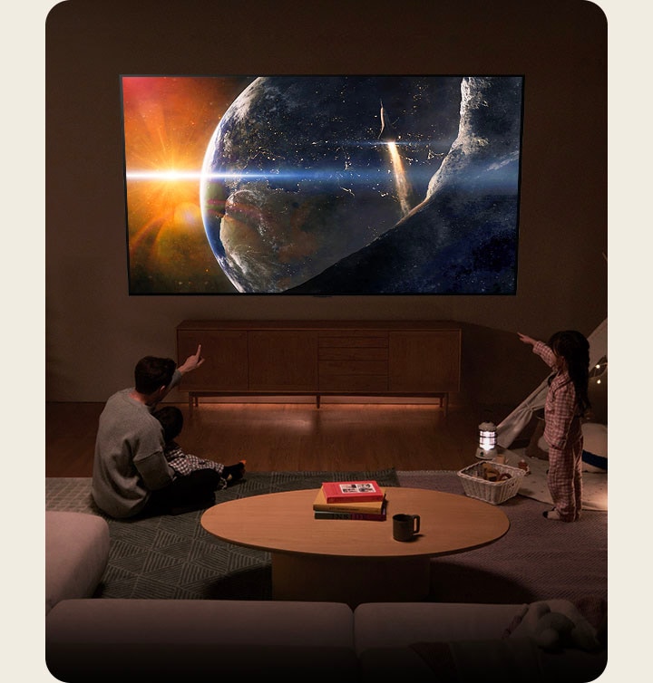 Una familia sentada en el suelo de un salón poco iluminado junto a una pequeña mesa, mirando hacia un televisor LG montado en la pared que muestra la Tierra desde el espacio.