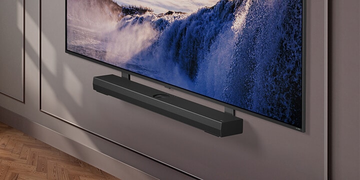 El soporte Synergy se muestra con LG QNED TV. El soporte Synergy y el televisor LG QNED se unen mientras se desplaza hacia abajo para mostrar la parte inferior del televisor. Aparece la barra de sonido LG, tal como está colocada en el soporte Synergy. El fondo aparece con la pared.