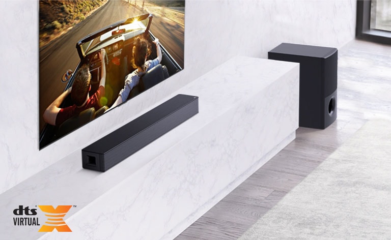 La TV está en la pared, la barra de sonido LG est a debajo en un estante de mármol blanco con un altavoz de graves a su derecha. La TV muestra una pareja en un auto.