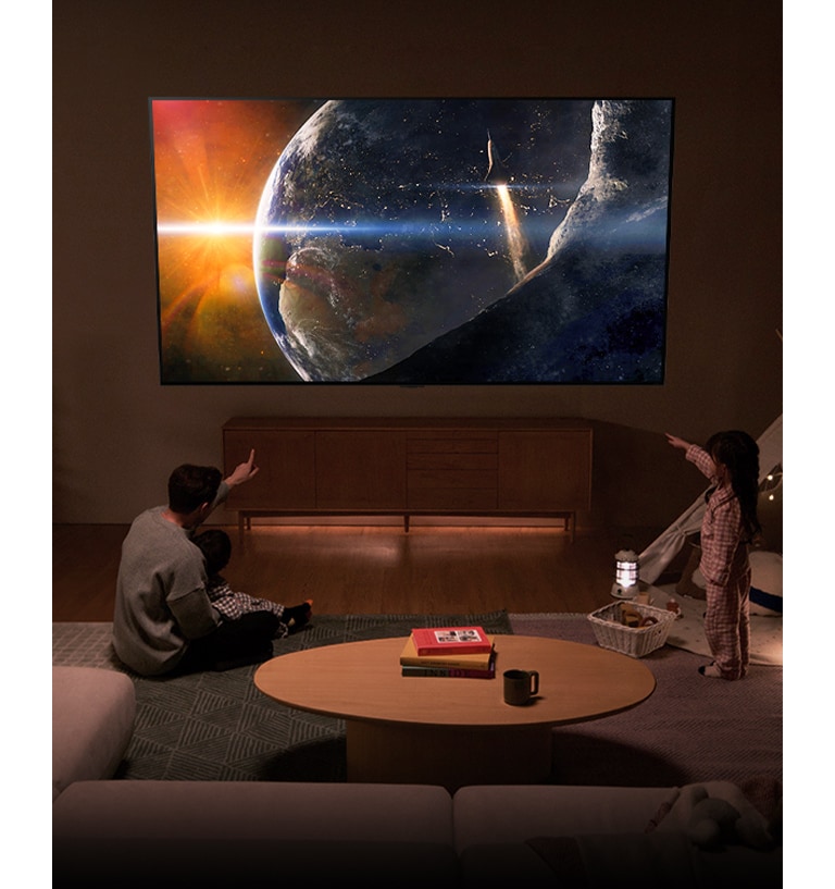 Una familia sentada en el suelo de un salón poco iluminado junto a una pequeña mesa, mirando hacia un televisor LG montado en la pared que muestra la Tierra desde el espacio.