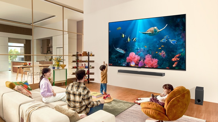 Una familia en una sala de estar con un televisor LG ultragrande montado en la pared, con una escena del océano que incluye corales y una tortuga en la pantalla.