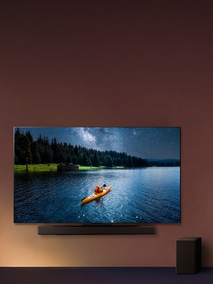 Televizor LG a soundbar připevněné na zdi a subwoofer na podlaze vpravo. Na televizoru je zobrazena osoba na kajaku na jezeře a na zdi se rozlévají jemné stíny.