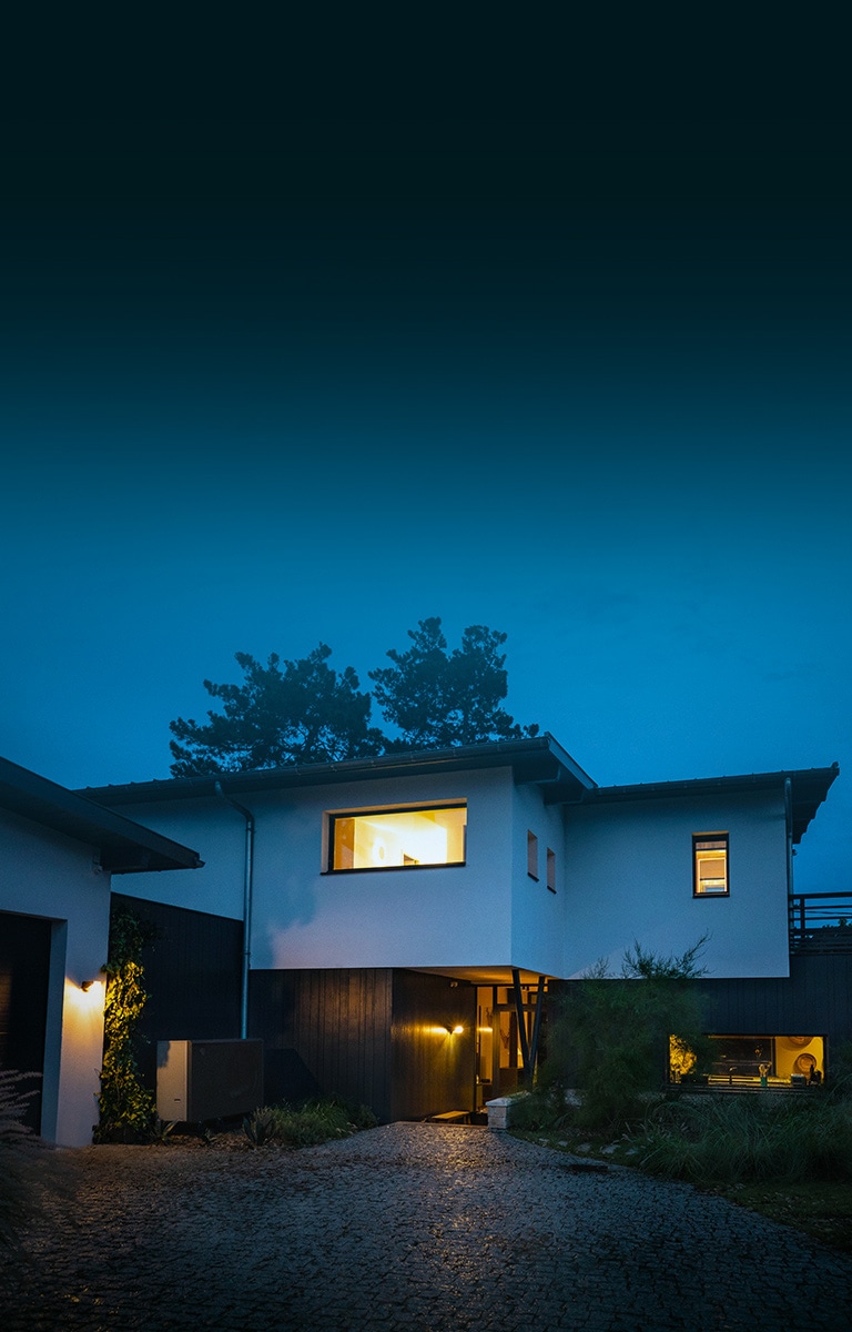 Celkový exteriér domu s rozsvícenými teplými světly za chladného zimního večera. Před domem je nové tepelné čerpadlo LG THERMA V typu vzduch-voda černé barvy. 