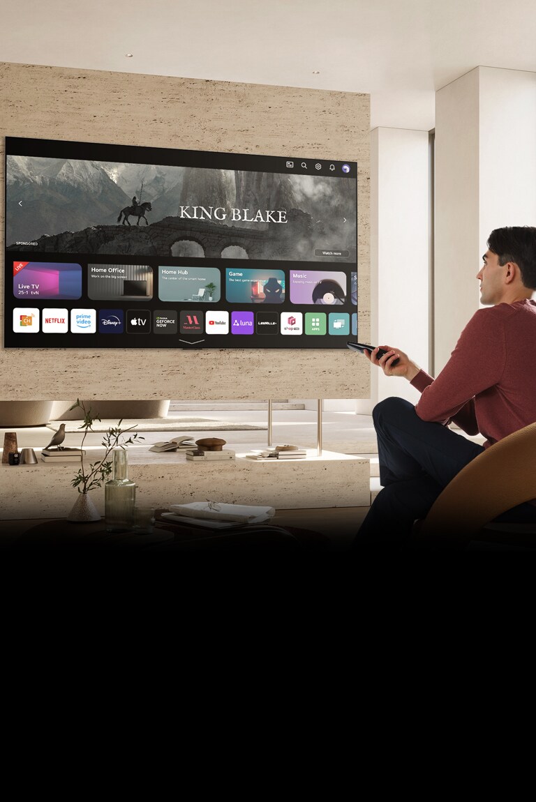Muž drží v pravé ruce dálkový ovladač a sleduje velký televizor naproti němu. Na televizní obrazovce se zobrazí „Zcela nová domovská obrazovka“.