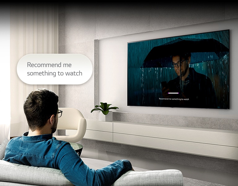 Muž sedí na pohovce čelem k televizoru. Nad jeho hlavou se vznáší bublina s textem „Recommend me something to watch“. Na obrazovce se přehrává video muže s deštníkem a zobrazuje se uživatelské rozhraní, které je rozpoznáno hlasem.