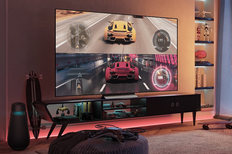 V červeně osvícené herní místnosti se nachází velká počítačová obrazovka, na které vidíme závodní hru s jedoucím žlutým a červeným autem. Vlevo je umístěn samostatný herní reproduktor a kolem něj je rozmístěno příslušenství pro hry a zařízení.