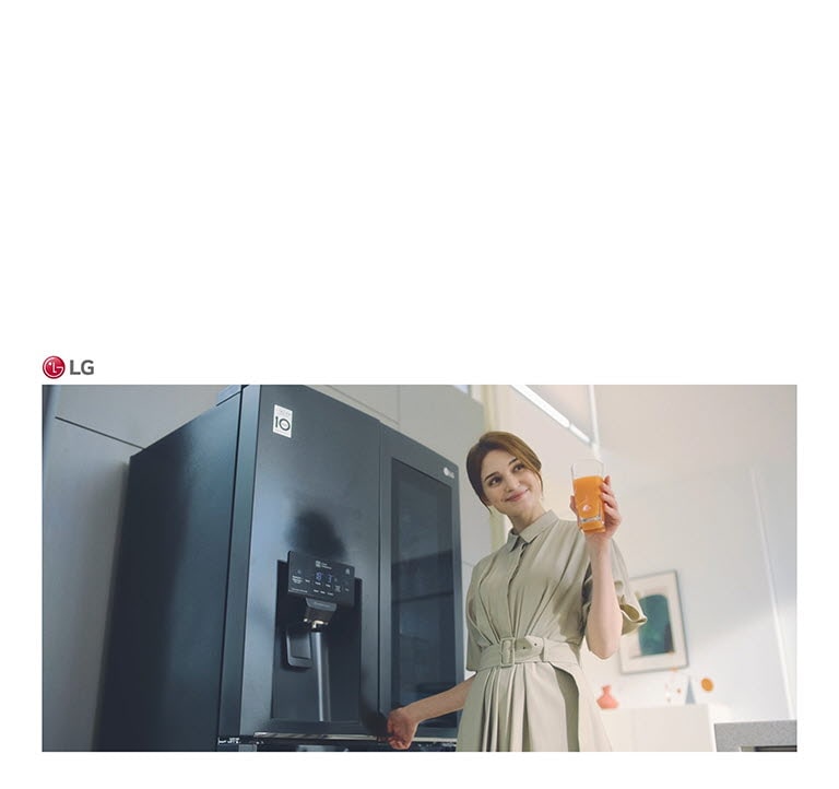 Žena stojí před chladničkou, drží sklenici džusu a usmívá se, když zavírá dveře.