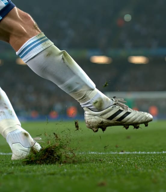 Ein Fußballspieler mit weißen Fußballschuhen und knielangen Stutzen stößt sich vom Rasen ab, wobei während des Spiels Erde und Gras aufgewirbelt werden.