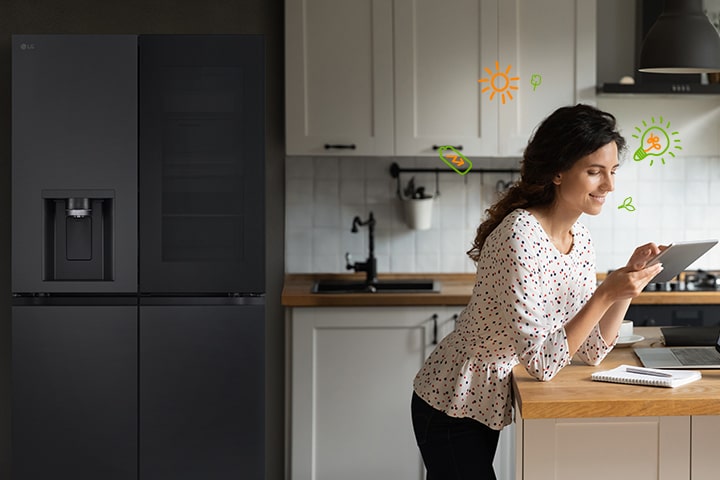 Vor dem Kühlschrank steht eine Frau, die lächelnd auf ihre Rechnung schaut; um die Frau herum sind Glühbirnen und Natursymbole zu sehen, die für Energieeffizienz stehen.