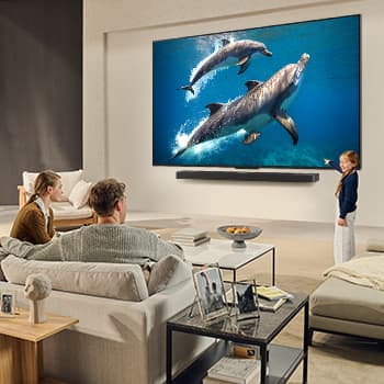 Eine Familie sitzt auf einer Couch vor einem LG QNED-Fernseher, der über einer LG Soundbar an der Wand montiert ist. Ein kleines Mädchen zeigt auf einen Bildschirm, auf dem zwei Delfine zu sehen sind. Der Titel spricht davon, dass jede Aktion auf einem großen Bildschirm noch spannender ist.