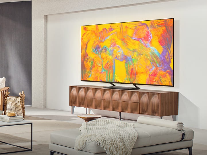 LG QNED TV steht über einer TV-Schublade in einem gemütlichen Wohnbereich. LG QNED TV zeigt einen abstrakten Hintergrund in Orange, Gelb, Lila und Rosa.