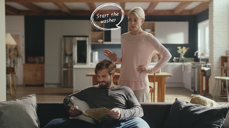 In einer modernen Smart-Küche verwendet eine Frau einen Sprachbefehl, um die Waschmaschine mit LG ThinQ AI zu starten, während ein Mann im Hintergrund auf dem Sofa liest.
