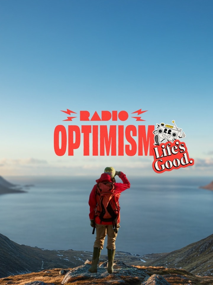 Eine Frau in einer roten Outdoor-Jacke steht an einem Ort, von dem aus man den weiten Ozean und schneebedeckte Berge sehen kann. Auf dem Bild sind außerdem das Logo mit dem Slogan „Radio Optimism“ sowie das Logo „Life’s Good.“ neben der Frau zu sehen.