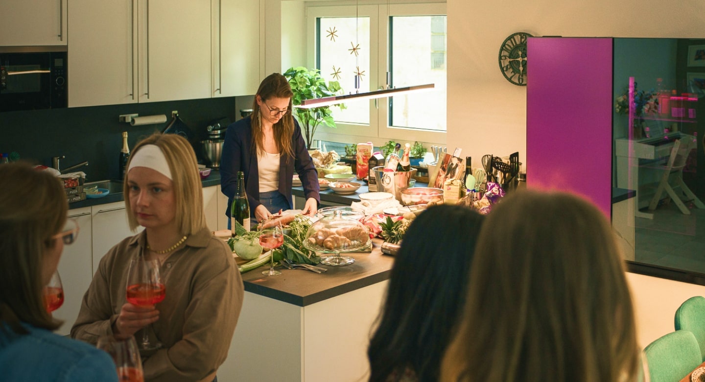 Auf der Hausparty unterhalten sich Freunde, während die Gastgeberin am Esstresen Essen vorbereitet.