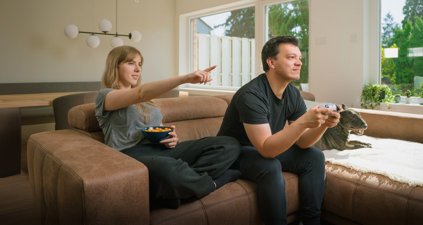 Ein Paar auf einer Couch, einer spielt ein Videospiel, der andere isst Snacks.